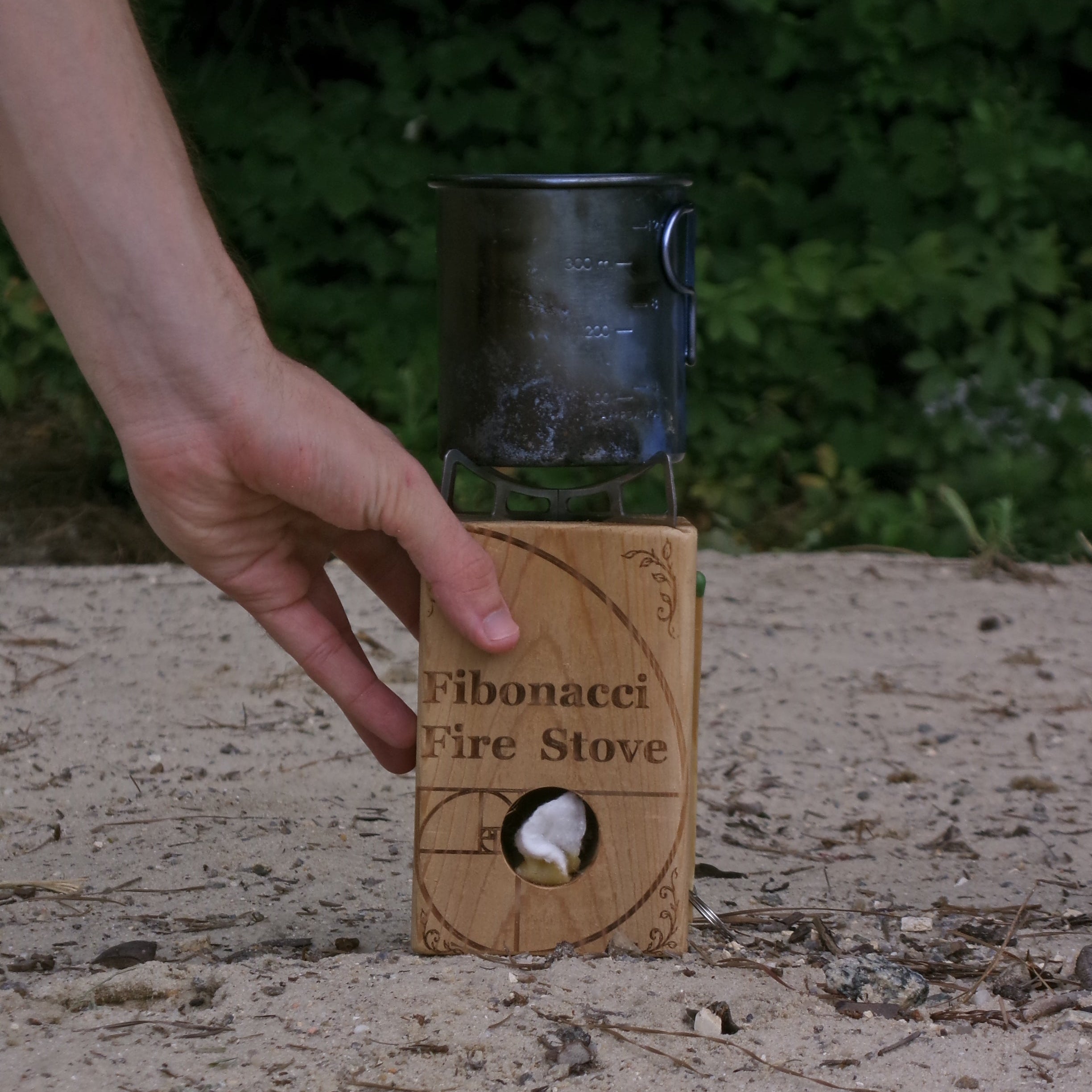 A hand holds a small wooden 'Fibonacci Fire Stove' outdoors on sandy ground with green foliage in the background. The stove has a circular cutout showing a white flame, and a dark metal pot sits on top.