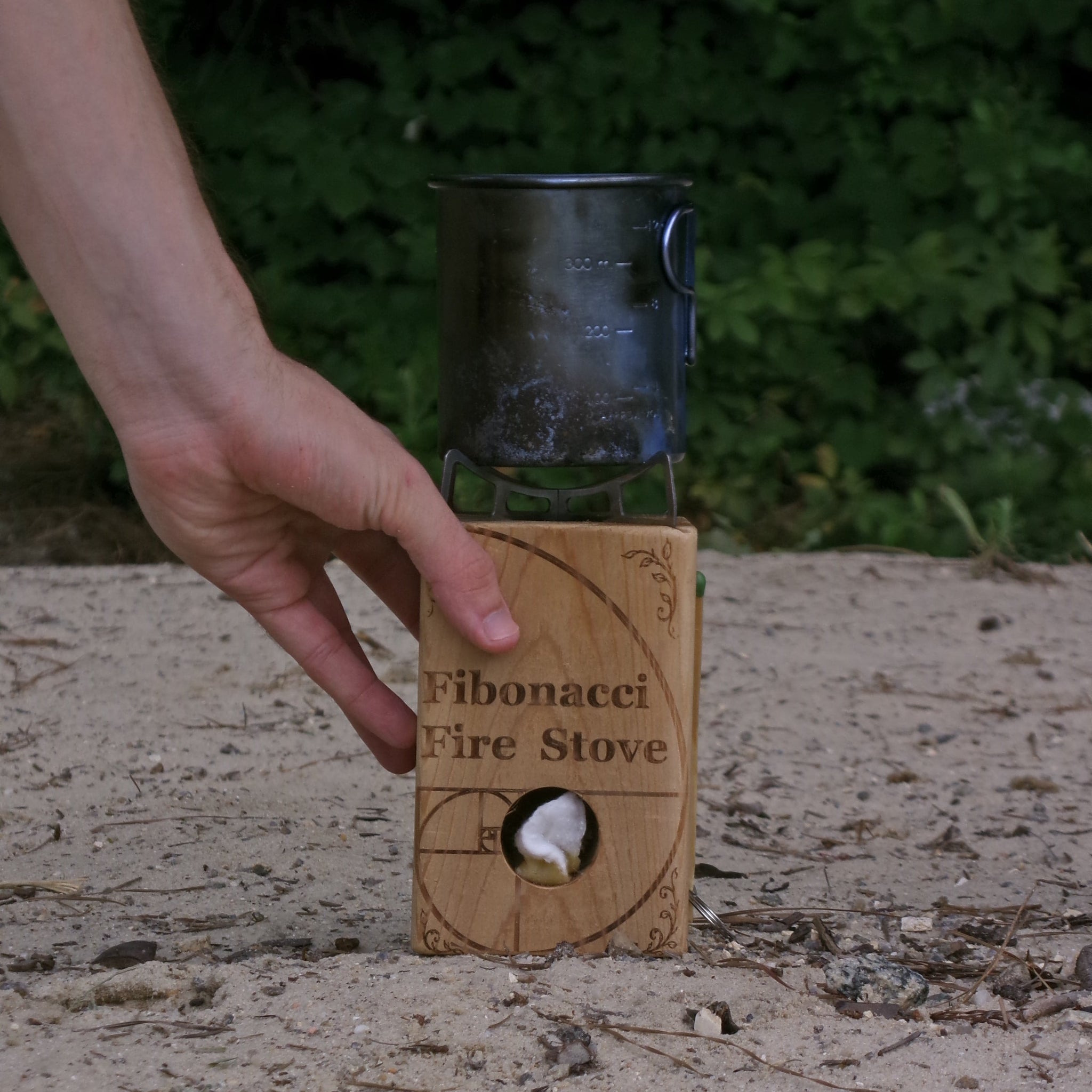 A hand holds a small wooden 'Fibonacci Fire Stove' outdoors on sandy ground with green foliage in the background. The stove has a circular cutout showing a white flame, and a dark metal pot sits on top.