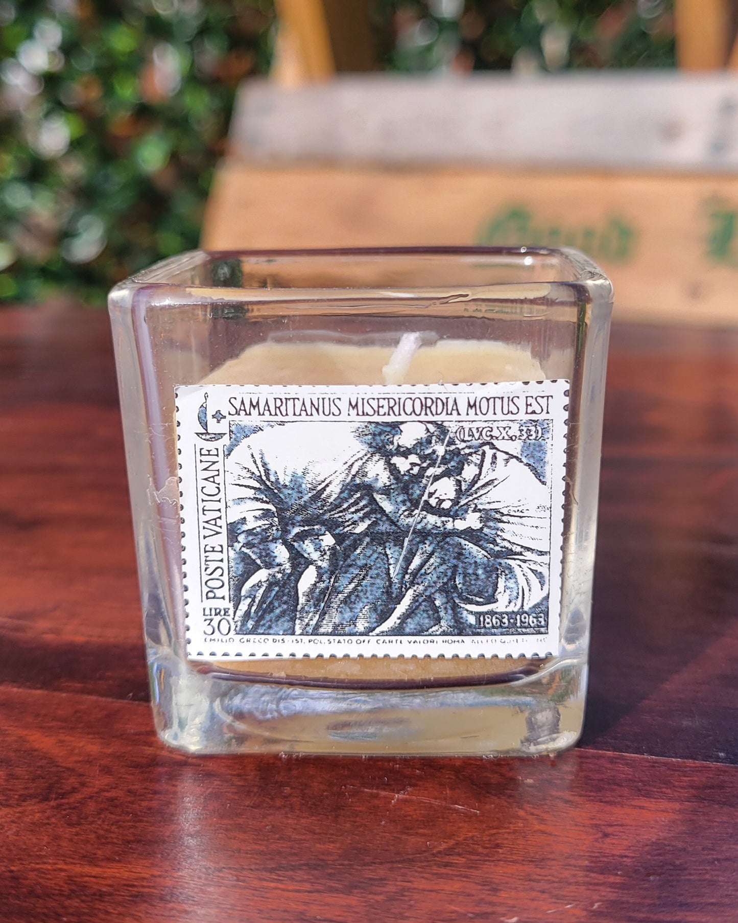 Glass candle on a coffee table featuring a stamp from the Vatican Postal Service depicting the parable of the Good Samaritan. The words "Samaritanus Misericordia Motus Est" are present on the stamp. This means "The Samaritan was moved to pity."
